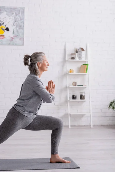 Mujer de mediana edad en guerrero con las manos orantes posan practicando yoga en la estera en casa - foto de stock