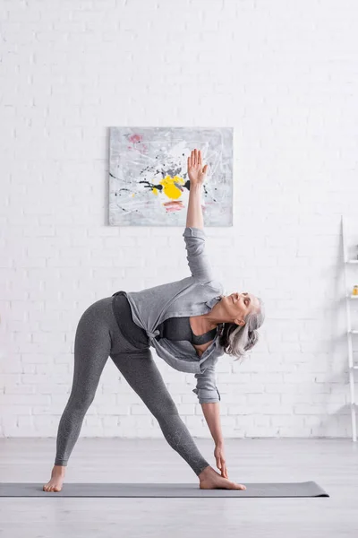 Frau mittleren Alters steht in Dreieck-Pose, während sie zu Hause Yoga auf Matte praktiziert — Stockfoto