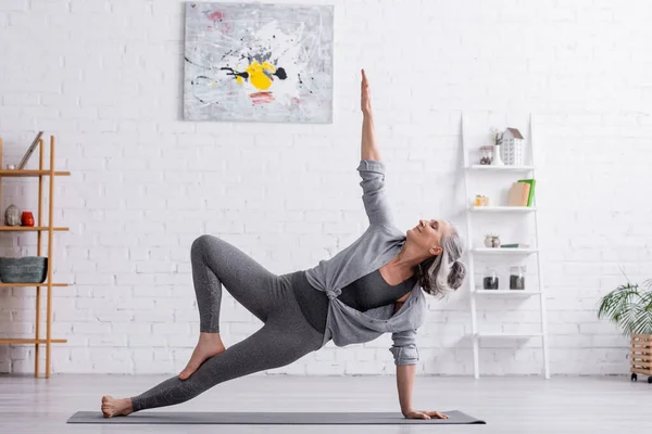 Mulher de meia-idade flexível em sportwear com mão levantada praticando ioga no tapete em casa — Fotografia de Stock