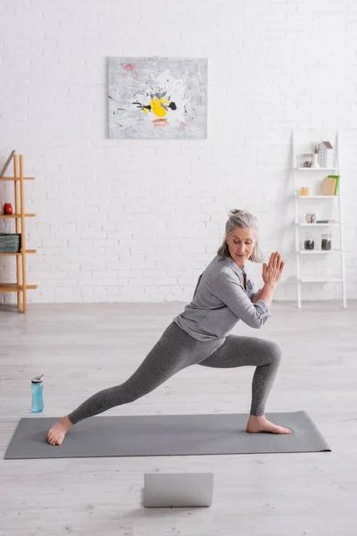 Femme d'âge moyen en posture guerrière avec des mains priantes pratiquant le yoga sur tapis près d'un ordinateur portable — Photo de stock