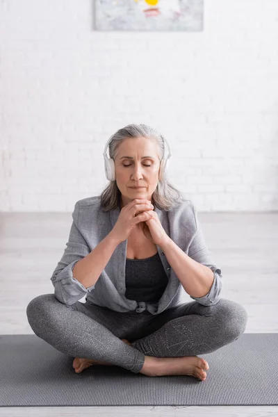 Mujer de mediana edad en auriculares inalámbricos meditando mientras está sentada en la pose de loto en la esterilla de yoga - foto de stock