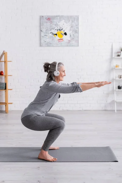 Side view of cheerful middle aged woman in sportwear and headphones doing squat on mat at home — Stock Photo