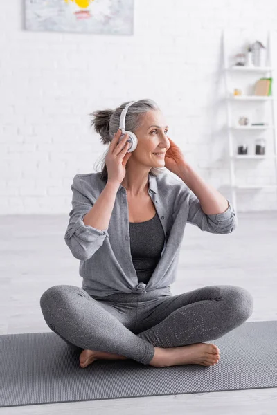 Happy middle aged woman in wireless headphones sitting in lotus pose on yoga mat at home — Stock Photo