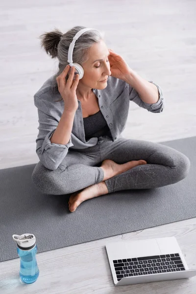 Hochwinkelaufnahme einer Frau mittleren Alters in drahtlosen Kopfhörern, die meditiert, während sie im Lotus auf einer Yogamatte in der Nähe des Laptops posiert — Stockfoto