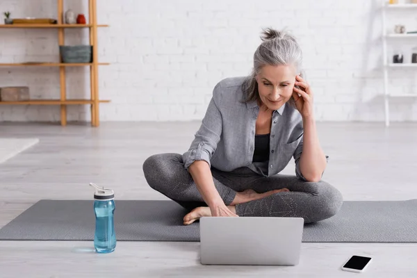 Middle aged woman in sportwear sitting in lotus pose on yoga mat and watching online tutorial on laptop — Stock Photo