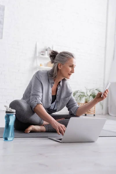 Middle aged woman sitting in lotus pose on yoga mat and looking at smartphone near laptop — Stock Photo