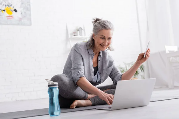 Feliz mulher de meia-idade sentado em pose de lótus no tapete de ioga e assistindo tutorial on-line no laptop e segurando smartphone — Fotografia de Stock