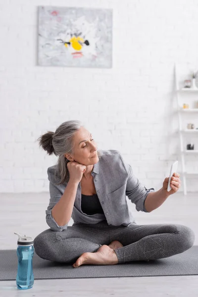 Middle aged woman sitting in lotus pose on yoga mat and using smartphone — Stock Photo