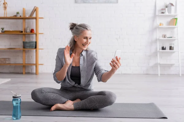 Mujer de mediana edad sentada en una pose de loto en una esterilla de yoga y saludando con la mano mientras recibe una videollamada en un teléfono inteligente - foto de stock