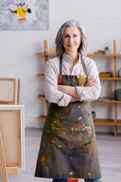 Joyful middle aged artist in apron standing with crossed arms near easel — Stock Photo