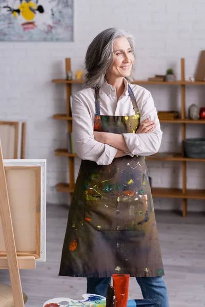 Cheerful middle aged artist in apron standing with crossed arms near easel — Stock Photo