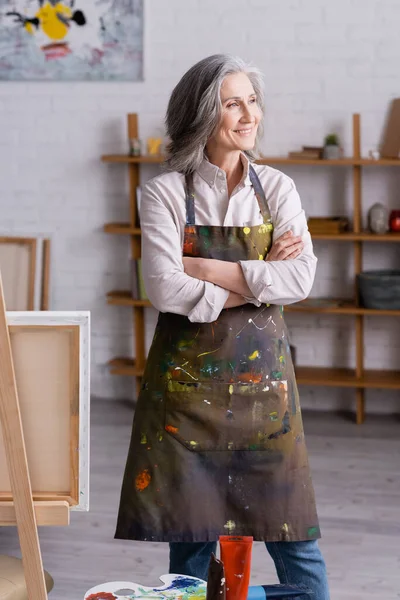 Fröhliche Künstlerin mittleren Alters in Schürze, die mit verschränkten Armen neben Staffelei steht — Stockfoto