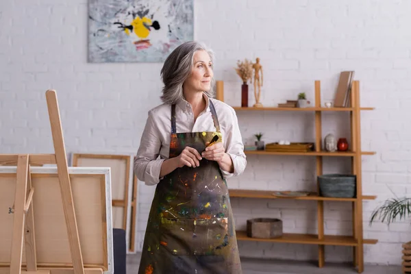 Middle aged artist in apron holding paintbrush near easel — Stock Photo