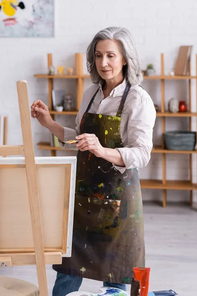 Middle aged artist in apron holding paintbrush while painting at home — Stock Photo