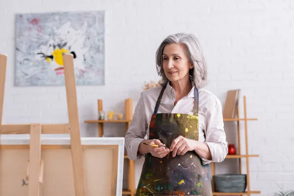 Pleased middle aged artist in apron with spills holding paintbrush and looking at canvas on easel — Stock Photo