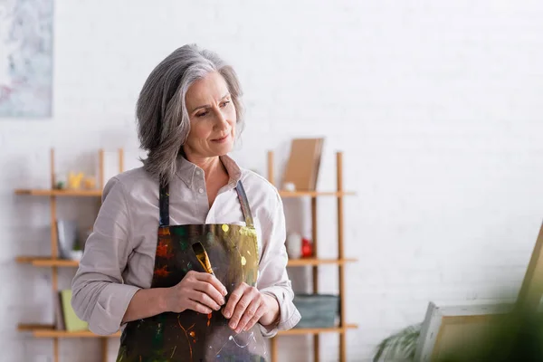 Artista de meia idade em avental com derramamentos segurando pincel em casa — Fotografia de Stock