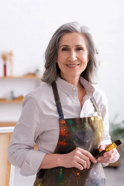 Cheerful middle aged artist in apron with spills holding paintbrush — Stock Photo