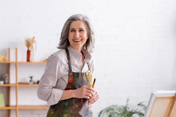 Alegre artista de mediana edad en delantal con derrames sosteniendo pincel cerca del caballete — Stock Photo