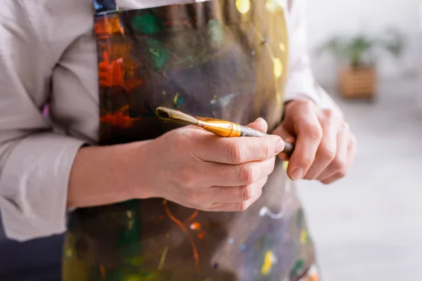 Partial view of middle aged artist in apron with spills holding paintbrush — Stock Photo