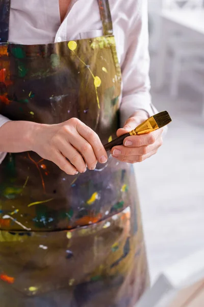 Cropped view of middle aged artist in apron with spills holding paintbrush — Stock Photo