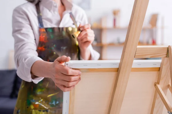 Cropped view of middle aged artist holding paintbrush near easel with canvas — Stock Photo