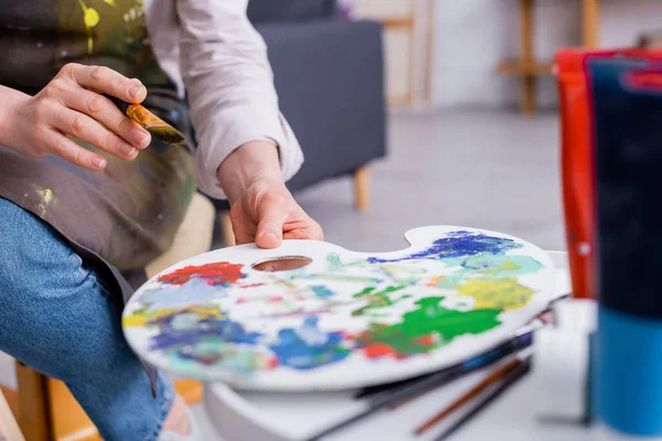 Cropped view of middle aged artist holding paintbrush and reaching palette with colorful paints — Stock Photo