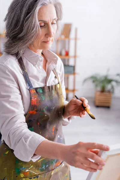 Middle aged woman holding paintbrush near easel with canvas — Stock Photo