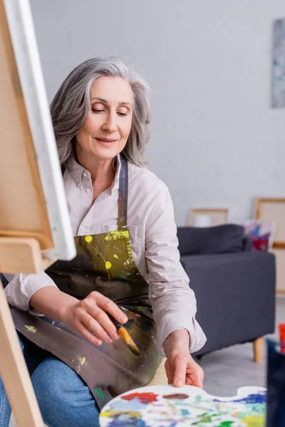 Artista de mediana edad sosteniendo pincel y llegar a la paleta con pinturas de colores - foto de stock