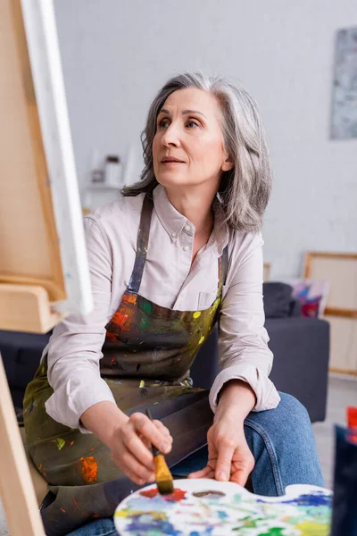 Artista de mediana edad sosteniendo pincel y paleta con pinturas de colores mientras mira la lona - foto de stock