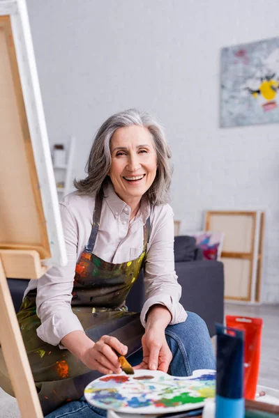 Alegre artista de mediana edad sosteniendo pincel y paleta con pinturas de colores cerca del caballete con lienzo - foto de stock