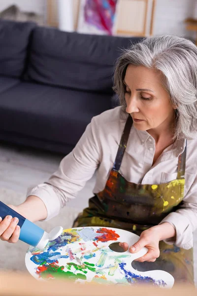 Frau mittleren Alters hält Tube mit blauer Farbe in der Nähe der Palette — Stockfoto