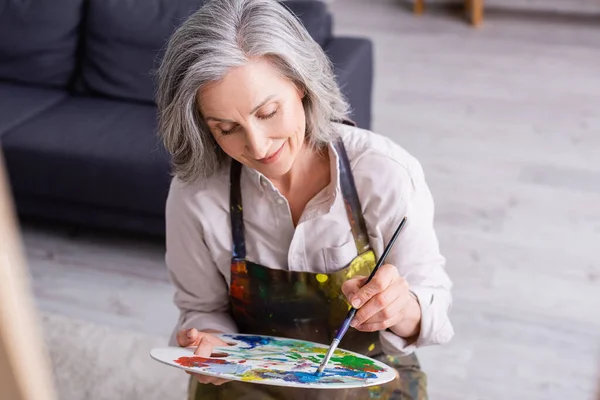 Piacere donna di mezza età in grembiule con pennello e tavolozza con vernici colorate — Foto stock
