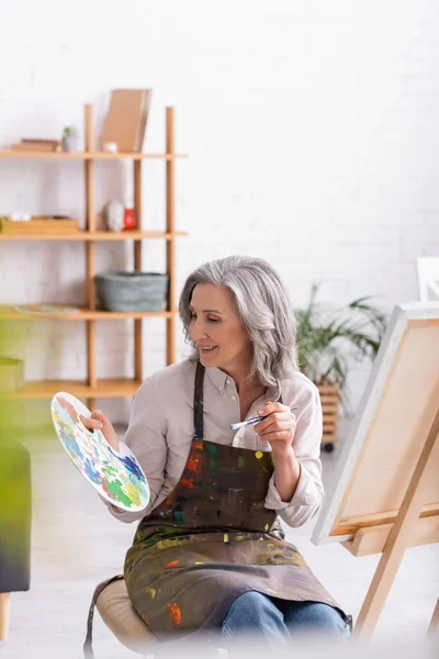 Cheerful mature woman in apron holding paintbrush and palette near easel at home with blurred foreground — Stock Photo