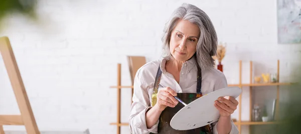 Pensive mature woman in apron holding paintbrush and palette while looking at easel, banner — Stock Photo