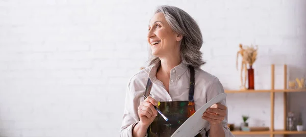 Smiling mature woman in apron holding paintbrush and palette while looking away, banner — Stock Photo