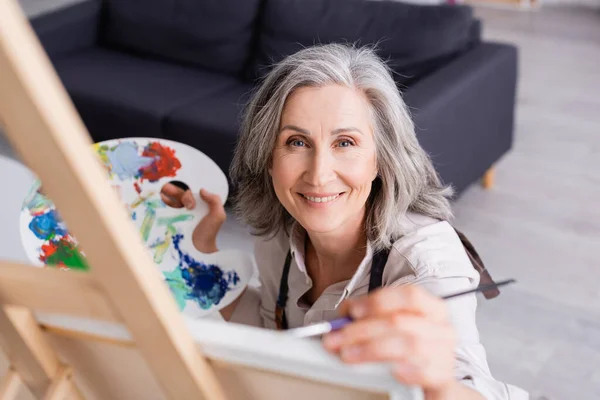 Sorrindo mulher madura segurando pincel e paleta enquanto pintura com primeiro plano borrado — Fotografia de Stock