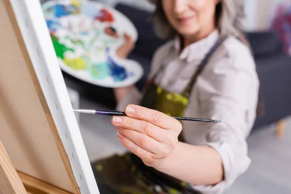 Partial view of mature woman holding paintbrush while painting on canvas with blurred foreground — Stock Photo