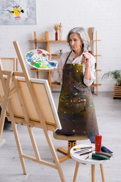 Зрілий художник тримає пензлик і палітру, стоячи біля полотна — стокове фото