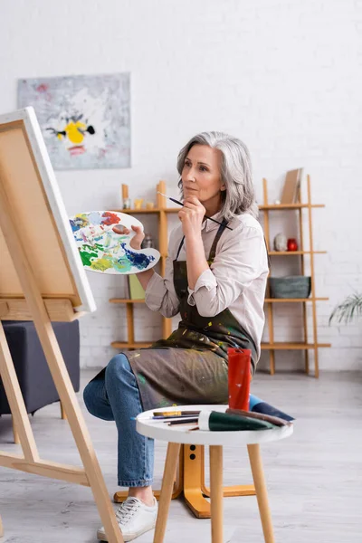 Artista maduro pensativo sosteniendo pincel y paleta mientras está sentado cerca de la lona - foto de stock