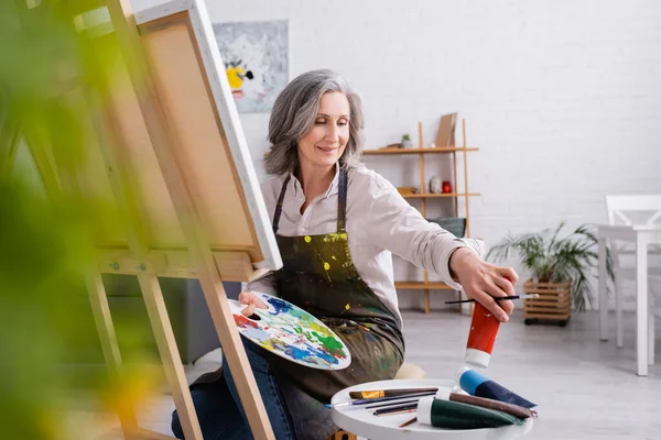 Mature woman holding paintbrush and palette while reaching red paint in tube and sitting near canvas — Stock Photo
