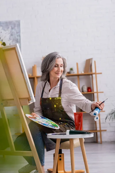 Pleased mature artist holding paintbrush and palette while reaching blue paint in tube and sitting near canvas — Stock Photo