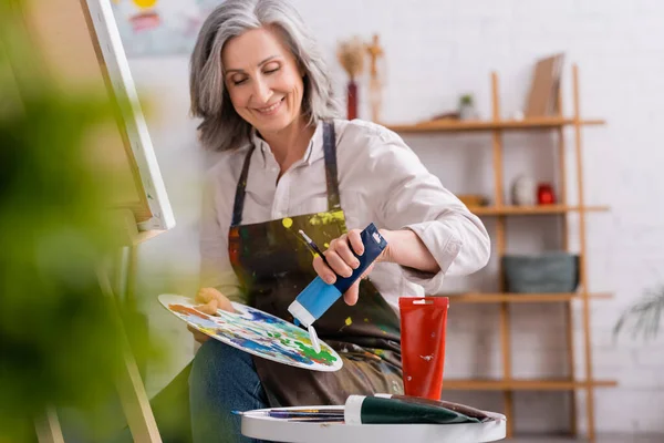 Pleased mature artist pouring blue paint on palette while sitting near canvas — Stock Photo