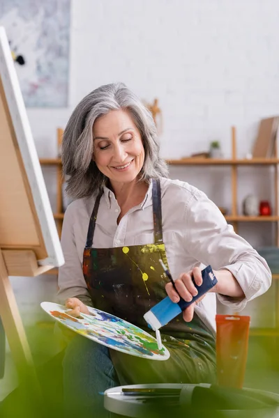 Glückliche reife Künstlerin gießt blaue Farbe auf Palette, während sie neben der Leinwand sitzt — Stockfoto