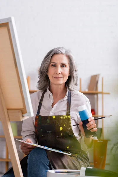 Mature artist holding tube with blue paint and palette while sitting near canvas — Stock Photo