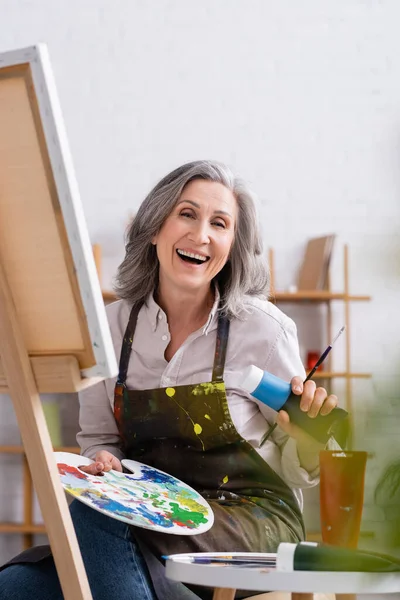 Happy mature artist holding tube with blue paint and palette while sitting near canvas — Stock Photo
