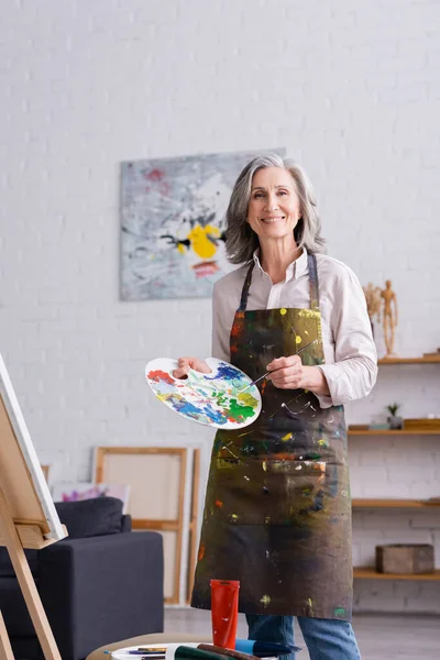 Positive middle aged woman holding paintbrush and palette with colorful paints near easel — Stock Photo
