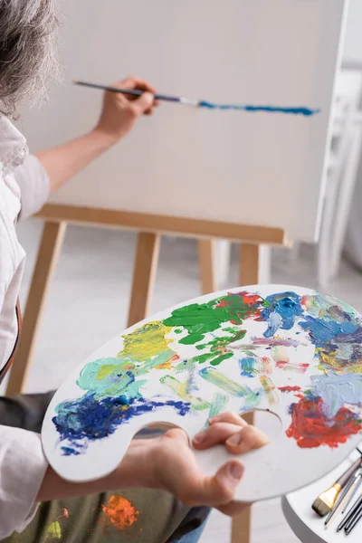 Cropped view of middle aged woman holding palette and paintbrush while painting on canvas — Stock Photo