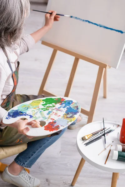 Mature woman holding palette and paintbrush and painting on canvas — Stock Photo