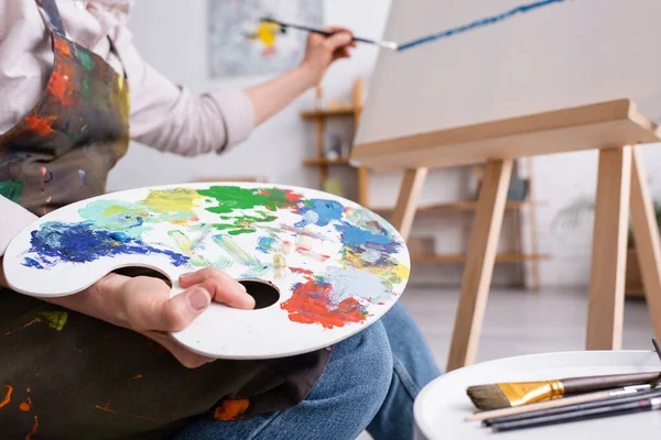 Cropped view of mature woman holding palette and paintbrush and painting on canvas — Stock Photo