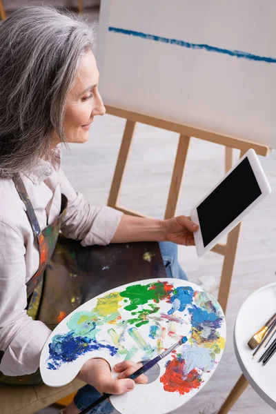 Зріла жінка тримає палітру і цифровий планшет з порожнім екраном біля полотна — стокове фото
